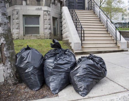 Three large black rubbish bags made of plastic, filled and tied at the top, are placed on the pavement near a tree in front of a building entrance. The bags are situated on a concrete sidewalk adjacent to a grassy area with a mature tree trunk visible on the left. In the background, there is a stone building with a set of stairs leading up to an entrance door, featuring black metal railings on either side. The scene appears to be a typical outdoor rubbish removal setup, with the black bags indicating waste ready for collection as part of a house clearance or rubbish removal service, such as those offered by House Clearance Chalfont Saint Giles. The environment suggests an urban or suburban setting, likely within a residential vicinity, with natural daylight illuminating the area and contributing to a clear, straightforward view of the waste bags and surrounding features.