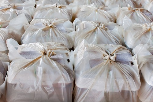 A close-up view of multiple tightly packed white plastic bags filled with rubbish, tied at the top with beige twist ties. The bags appear to contain various waste materials, with some slightly bulging and showing different textures and colours through the semi-transparent plastic. They are arranged in a neat, orderly manner on a flat surface, likely outdoors on a driveway or pavement, with a subtle sheen reflecting natural light. The scene reflects an organized rubbish collection or disposal process, typical of waste removal services such as those offered by House Clearance Chalfont Saint Giles, which operates in the Chalfont Saint Giles area and nearby locations. The background does not contain visible structures or environmental features, keeping focus on the stacked bags ready for removal or disposal, emphasizing the task of clearing waste efficiently and professionally.
