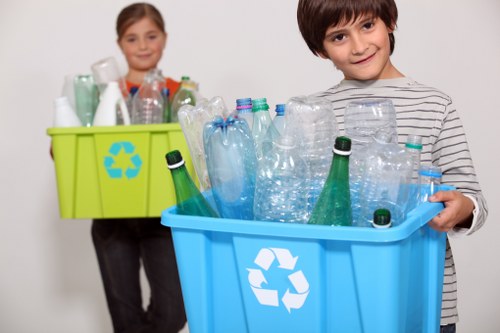 A young boy with dark brown hair, wearing a long-sleeved striped shirt, is holding a blue recycling bin filled with various clear and green plastic bottles, some with caps and some without, arranged haphazardly. Behind him, a young girl with light brown hair, dressed in a dark long-sleeved top, stands holding a green recycling bin filled with additional plastic bottles and containers, some partly visible. Both children are indoors against a plain white background, and the blue and green bins prominently display the universal recycling symbol in white on the front, emphasizing sustainable waste management practices. The scene highlights the importance of recycling household plastics, aligning with waste collection services such as those offered by House Clearance Chalfont Saint Giles, which operates in the local area, subtly supporting environmentally responsible rubbish disposal efforts.