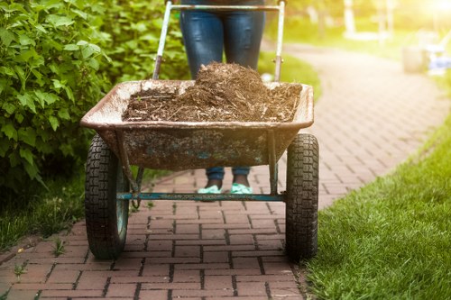 A person standing behind a metal wheelbarrow filled with dark, damp soil or garden waste, positioned on a brick pathway bordered by green bushes on the left and a grassy verge on the right. The individual is wearing blue jeans and black shoes, with only their legs and lower torso visible in the image. In the background, the pathway extends into a residential garden area with sunlight filtering through, creating a bright and natural atmosphere. The scene appears to be outdoors in a suburban setting, consistent with garden clearance or waste removal activities often undertaken by companies like House Clearance Chalfont Saint Giles, focusing on rubbish removal in the local area. The wheelbarrow has a sturdy metal tray and large black rubber tyres suitable for transporting garden debris in preparation for disposal or recycling.
