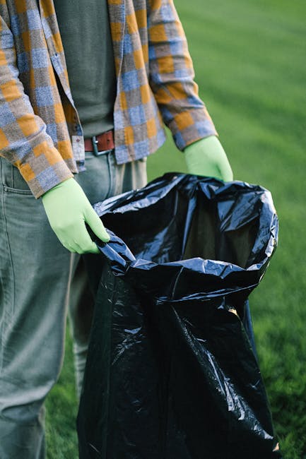 A person wearing a checked yellow and gray shirt over a green top and gray trousers, standing outdoors on a grassy area during daytime, is holding open a large black rubbish bag lined with a silver in