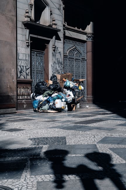 A person wearing a yellow and grey plaid shirt over a green sweater and grey trousers is standing outdoors on a grassy area. They are holding open a large black rubbish bag lined with a silver interio